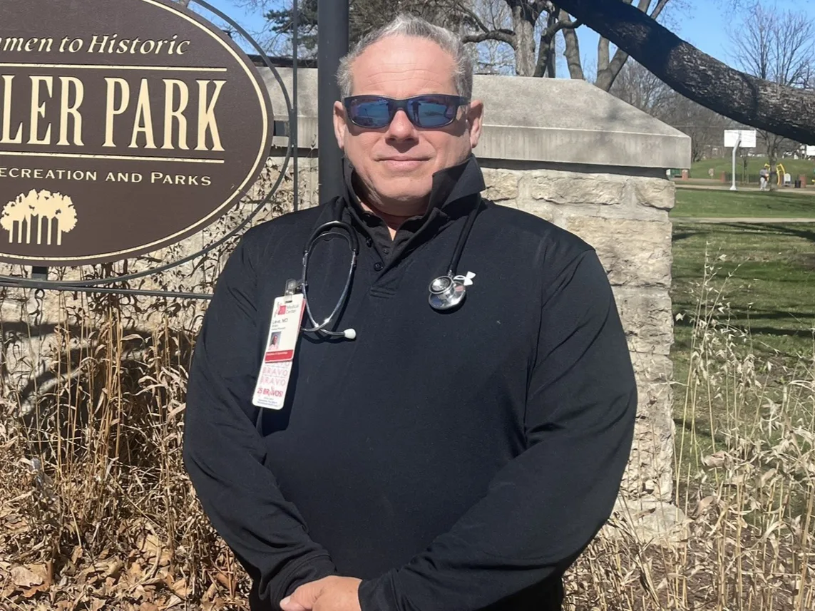 Dr Leve in clinic blacks with a stethoscope, beside the Schiller Park sign in German Village.